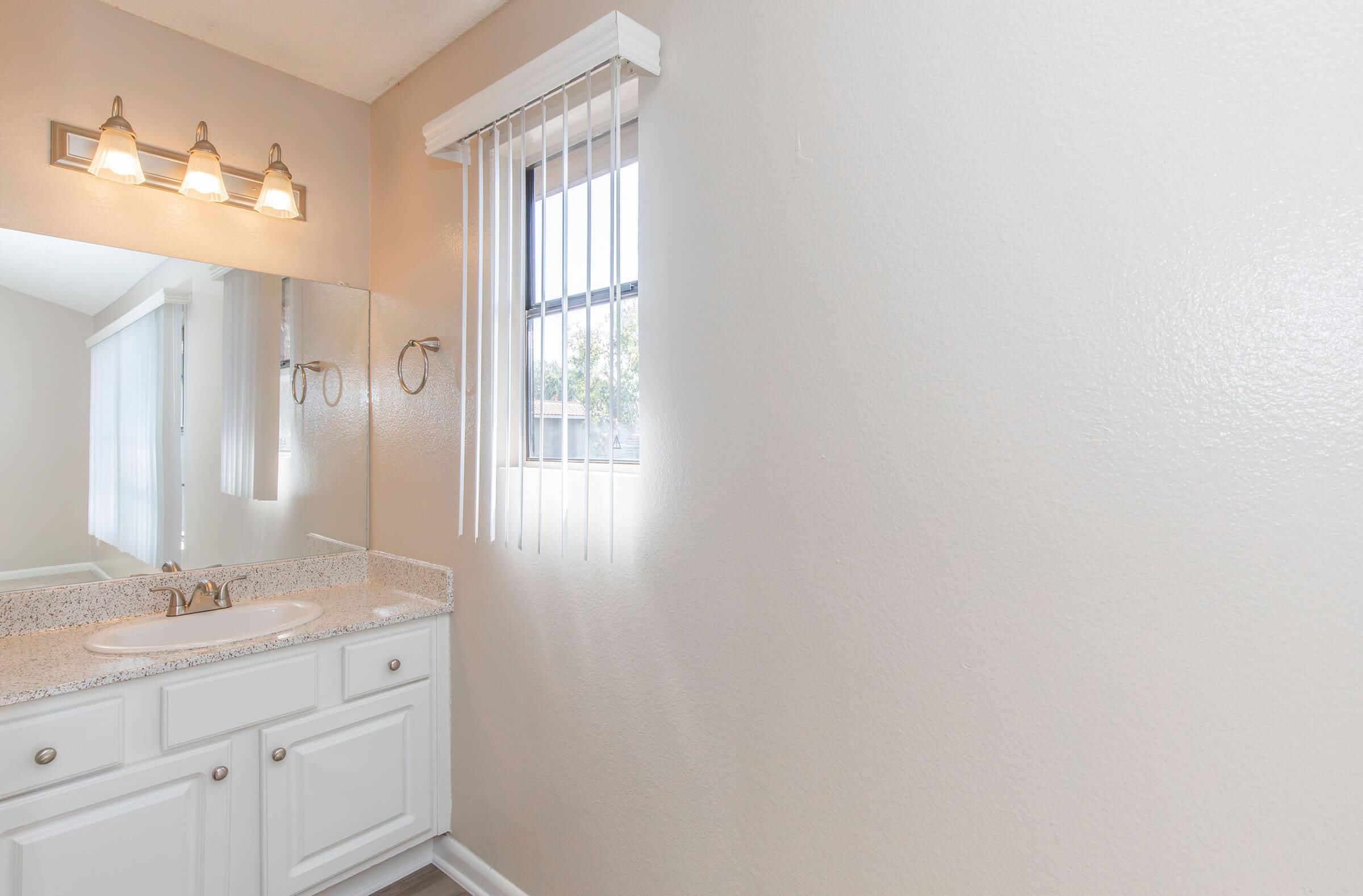 A clean bathroom featuring a granite countertop with a sink, white cabinets, and a large mirror. Natural light enters through a window with vertical blinds. The walls are painted a neutral color, and the lighting fixtures above the mirror provide warm illumination.
