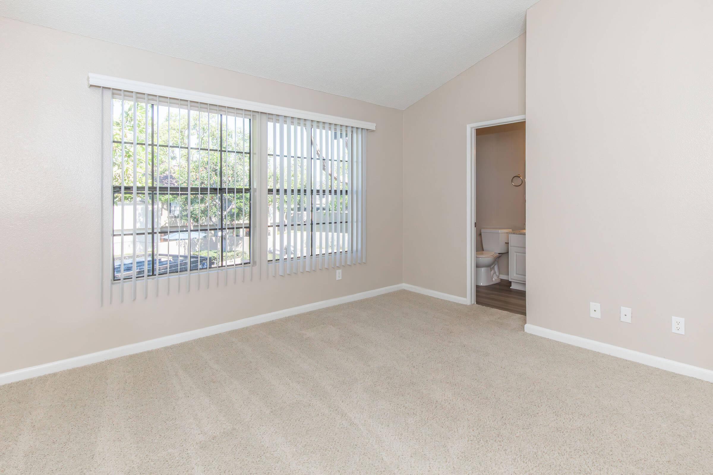 A spacious, empty room featuring beige walls and carpet. Large vertical blinds cover a window, allowing natural light to enter. A doorway on the right leads to a bathroom area, while the overall ambiance is clean and inviting, ideal for personalization or decoration.