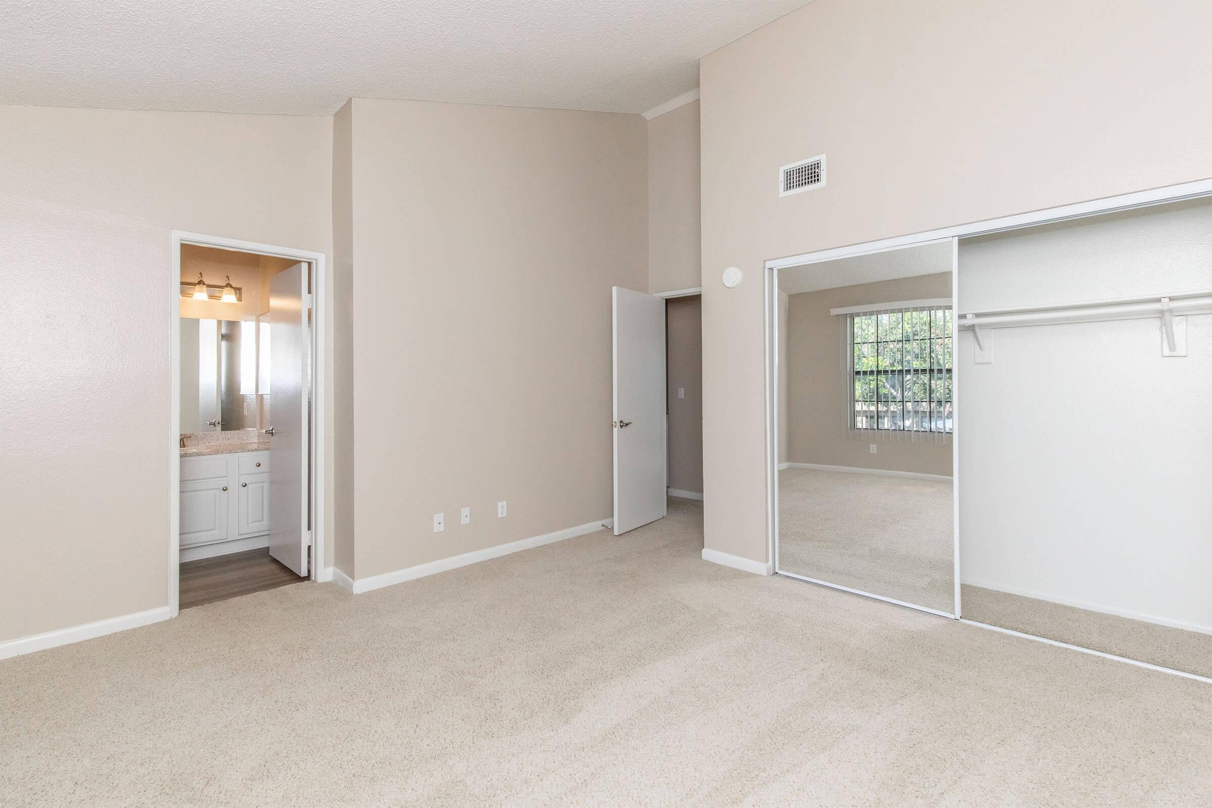 A spacious room with beige walls and carpeted flooring. There is a doorway leading to a bathroom with a vanity and mirror visible. A large mirrored closet is to the right, reflecting natural light from a window. The room features high ceilings and a neutral color palette, creating a bright and airy atmosphere.