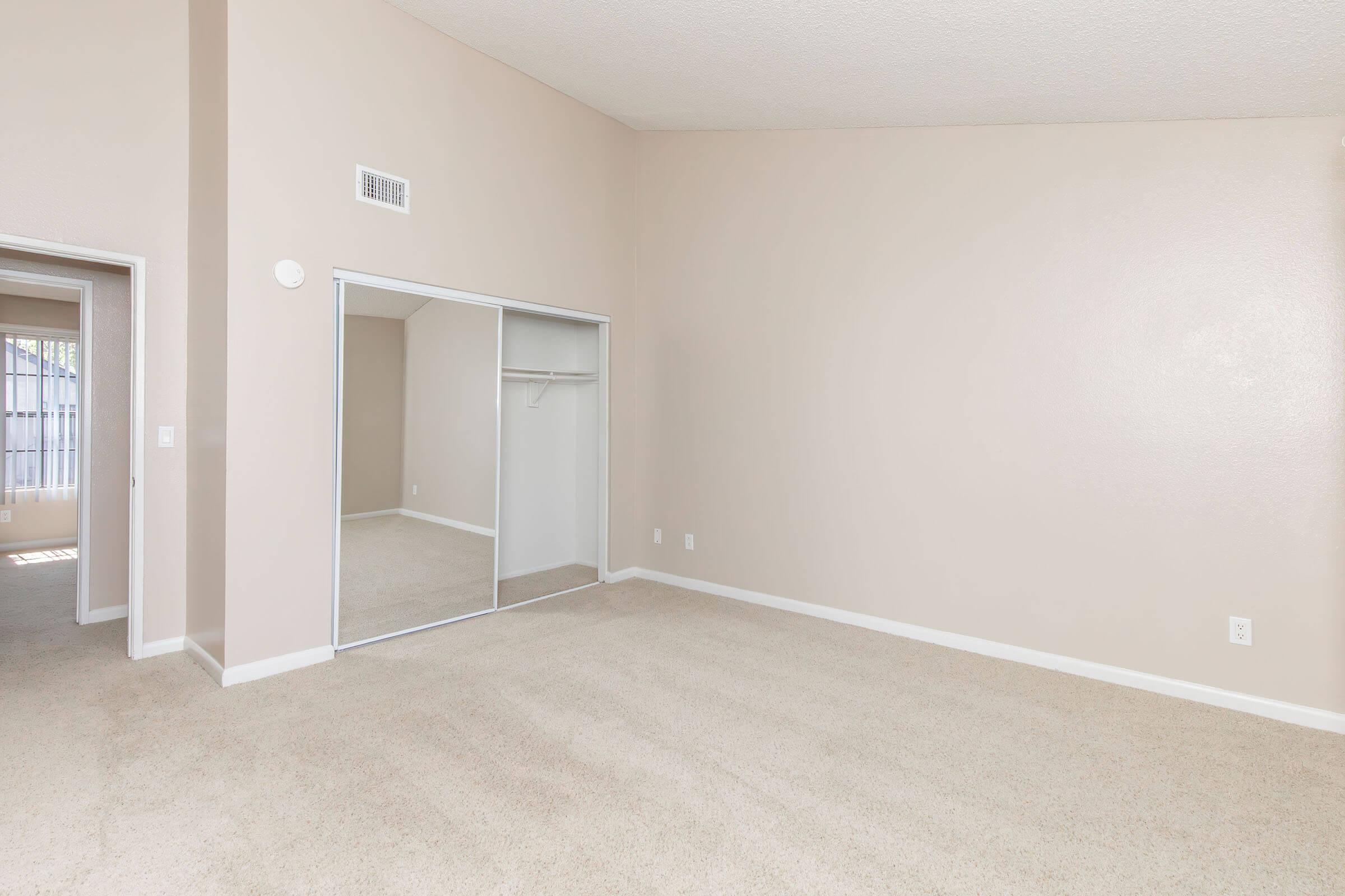 Interior view of a spacious, empty room with light beige walls and carpeted flooring. There is a large closet with mirrored sliding doors on one side, and a doorway leading to another room or hallway. The space is well-lit, showcasing a high ceiling and neutral decor, making it versatile for various furnishings.