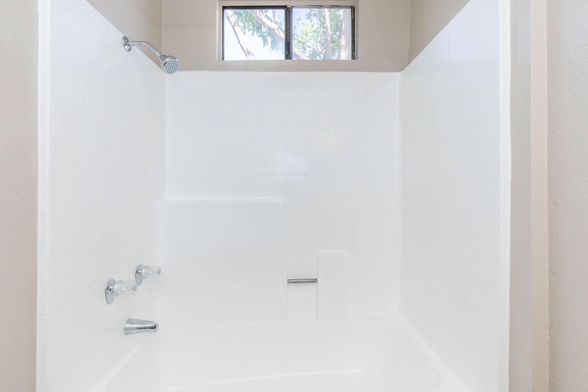 A clean, simple shower stall featuring white tiled walls and a showerhead. A small ledge is visible, and there is a window allowing natural light to enter the space. The overall aesthetic is bright and minimalist, emphasizing a fresh and functional design.