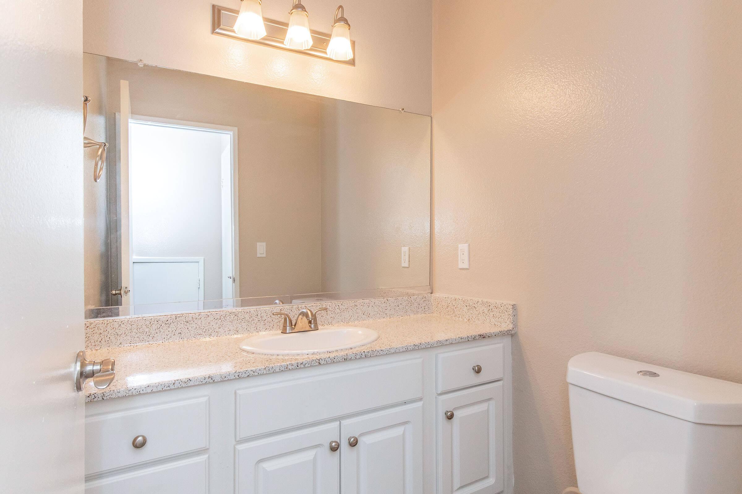 A well-lit bathroom featuring a large mirror above a granite countertop with a sink, accompanied by a white cabinet below. To the right, a white toilet is visible. The walls are a soft beige color, and there is a door leading to another room in the background.