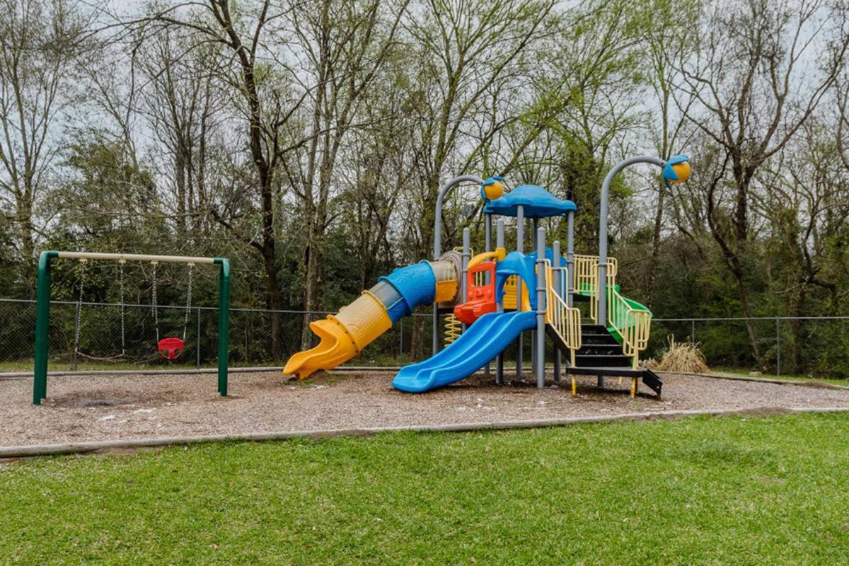 A colorful playground featuring slides in blue and yellow, climbing structures, and swings. The swings include a red seat for children. Surrounding the playground are trees and a grassy area, creating a natural setting for outdoor play.