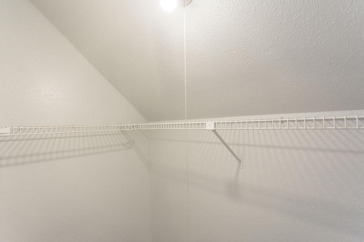 A bright, empty closet with white walls and a wire shelf. The ceiling is sloped, and there is a single overhead light fixture illuminating the space. The floor is not visible, and the overall atmosphere is clean and minimalistic.