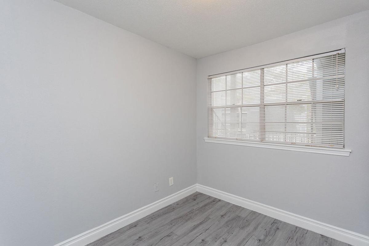 Empty room with light gray walls and a wooden laminate floor. A window with white blinds allows natural light to enter. The space is uncluttered, making it suitable for various uses, such as a home office or guest room.