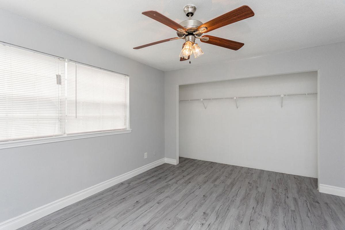 A bright, empty room featuring a ceiling fan with a wooden blade, light fixtures, and large window with blinds. The walls are painted light gray, and the flooring is a light wood laminate. There is a closet space with a hanging rod on one side, emphasizing the room's spaciousness.