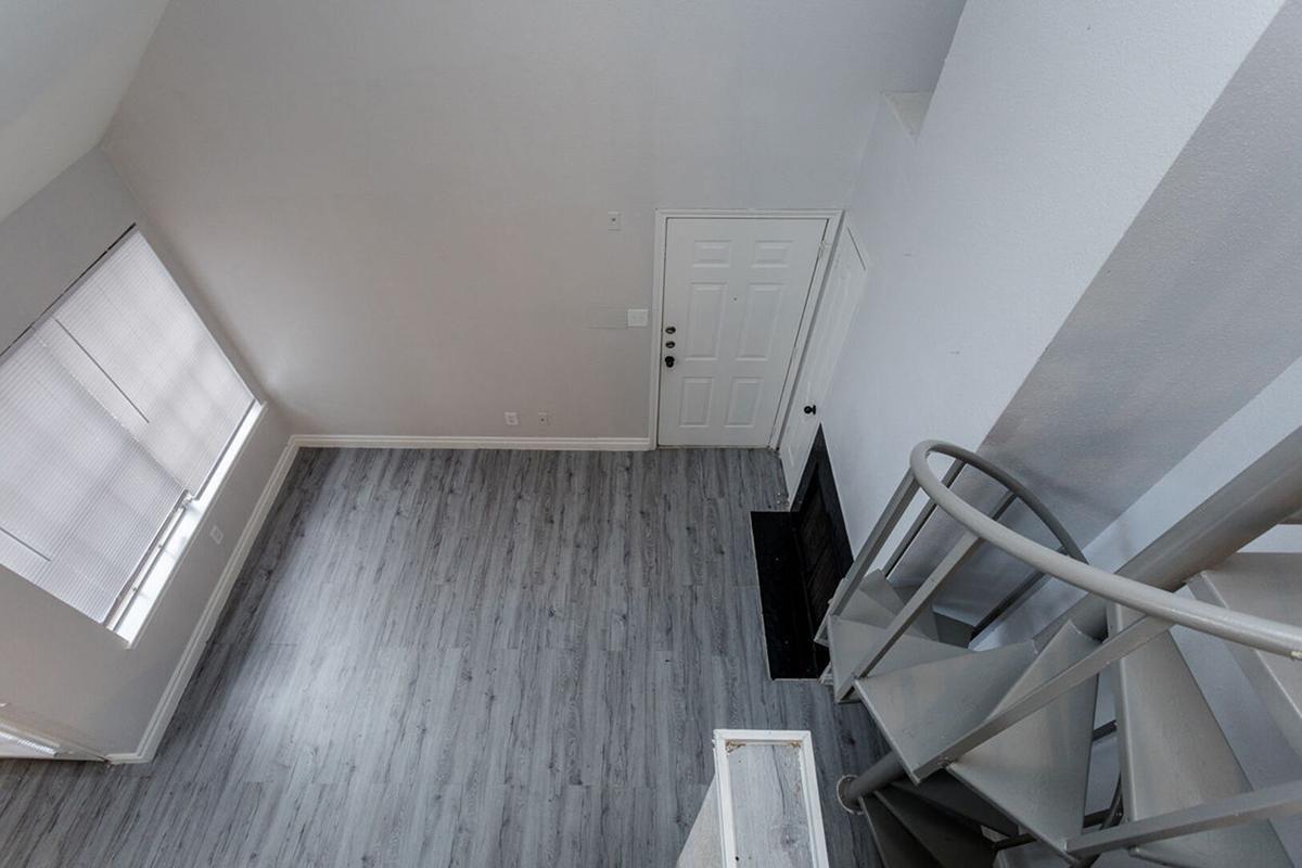 Aerial view of an empty, modern interior space with light grey walls and flooring. There's a staircase leading to an upper level, a front door on the right, and a large window allowing natural light. The room is minimalistic, giving it a spacious feel.