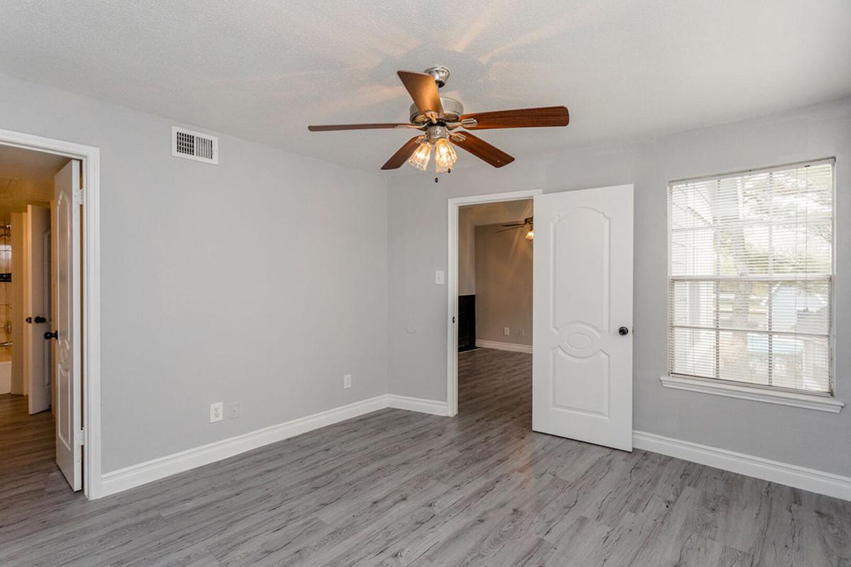 Interior view of a well-lit, empty bedroom featuring light gray walls and wood-like flooring. A ceiling fan with lights is centered in the room. There are two doorways: one leads to a bathroom and the other opens to an adjacent area. A window allows natural light to enter, enhancing the spacious feel of the room.