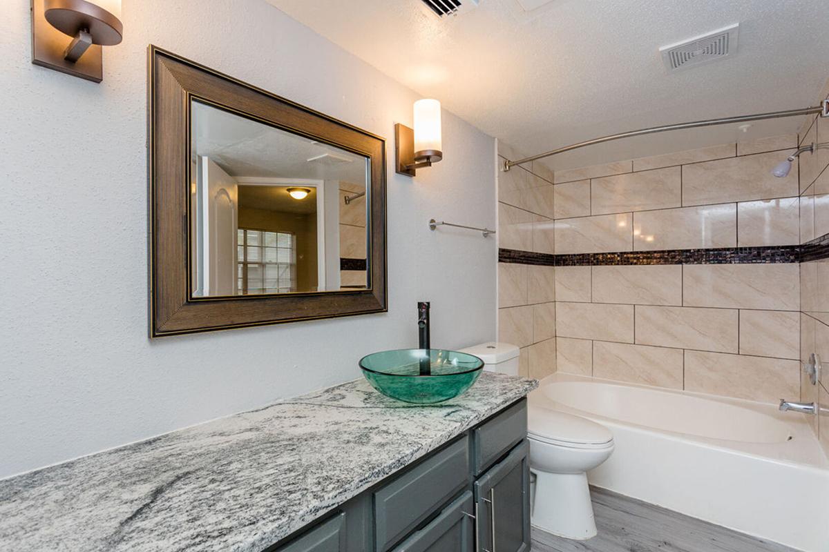 A modern bathroom featuring a granite countertop with a round glass sink, a large mirror above the sink, and a bathtub with a tiled surround. Soft lighting is provided by wall sconces, and a neutral color palette enhances the contemporary design.
