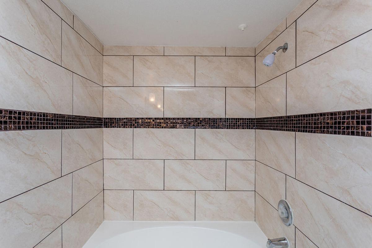 A tiled shower wall featuring light-colored tiles with a decorative dark mosaic strip running horizontally. The lower part has a bathtub edge visible. The overall design is modern and clean, ideal for a bathroom space.