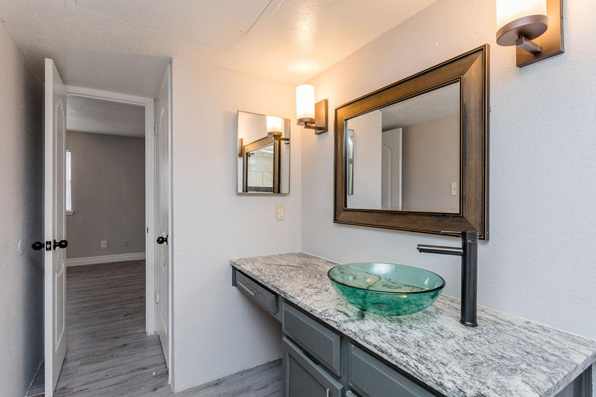 A modern bathroom featuring a granite countertop with a stylish green glass sink. Above the sink are two mirrors and wall-mounted light fixtures. The room has light gray walls and a door leading to another room in the background, showcasing a neutral decor style.