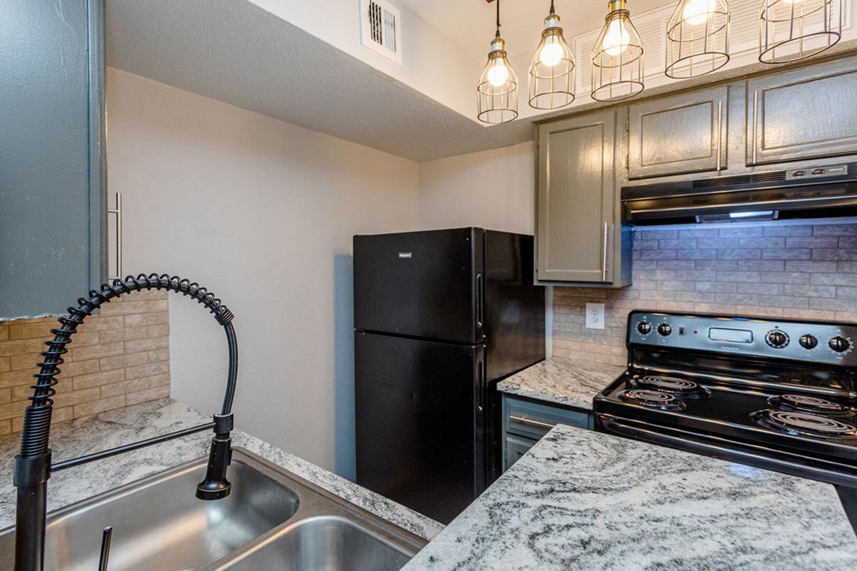 A modern kitchen featuring granite countertops, a stainless steel sink with a pull-down faucet, a black refrigerator, and an oven with a stovetop. Pendant lights hang above the counter, adding warm illumination to the space, which has a neutral-colored wall and cabinetry.