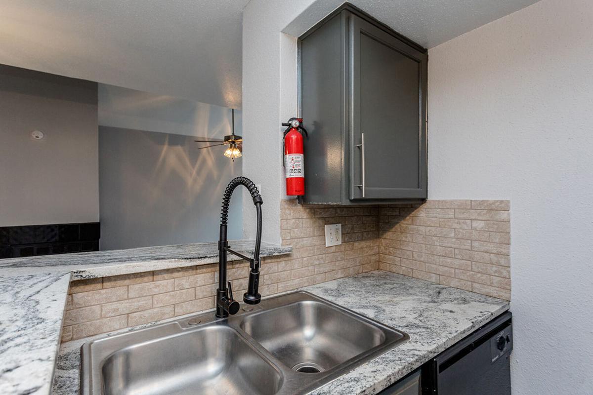 Modern kitchen featuring a double sink with a stylish black faucet, granite countertops, and a beige tiled backsplash. A fire extinguisher is mounted on the wall above the sink. The room is well-lit with a ceiling fan visible in the background, suggesting a functional and contemporary living space.