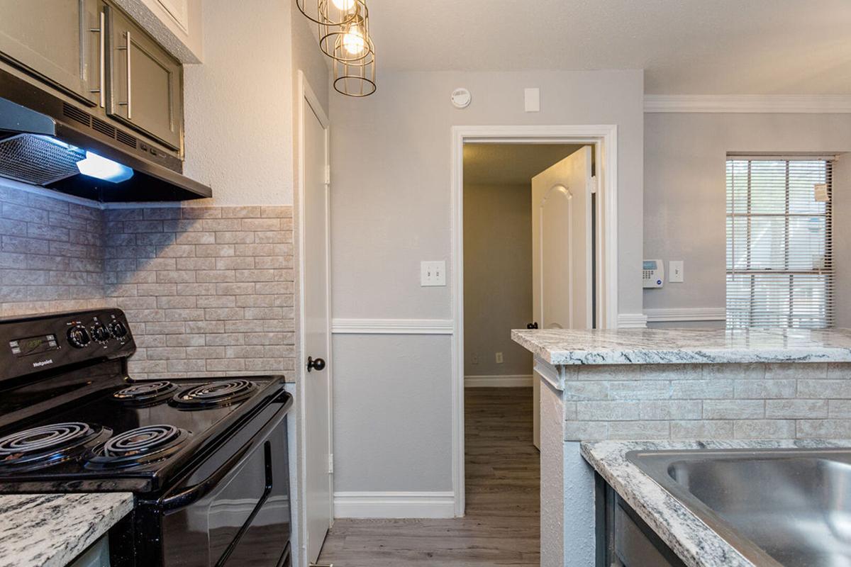 A modern kitchen featuring a black stove, granite countertops, and pendant lighting. The walls are light gray with a textured backsplash. A doorway leads to another room, and there are windows providing natural light. The overall design is sleek and contemporary.