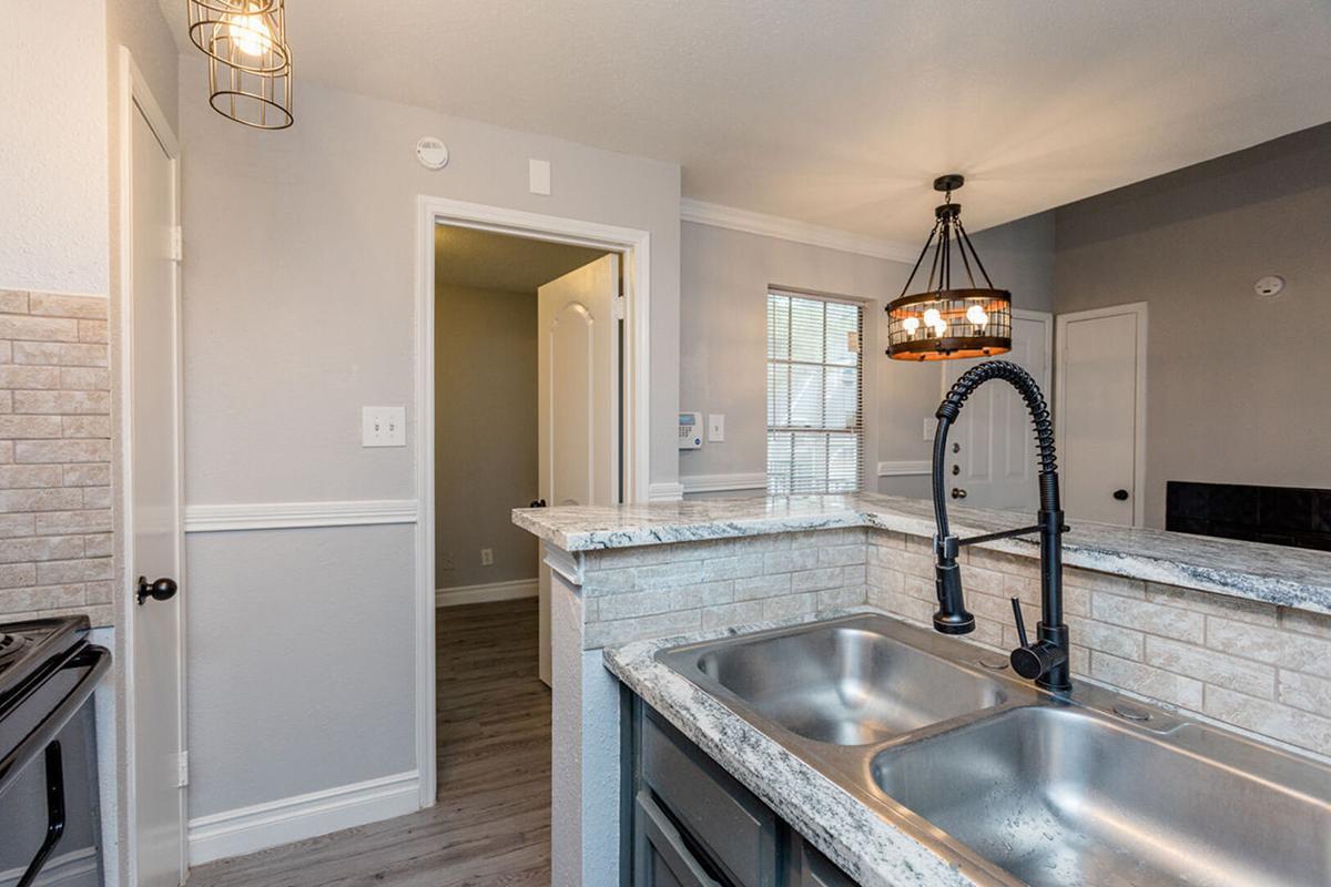 A modern kitchen with a double sink featuring a sleek faucet, granite countertops, and a stylish light fixture. The walls are painted in soft gray, and there's a doorway leading to another room. Natural light streams in through a window, enhancing the open space feel.