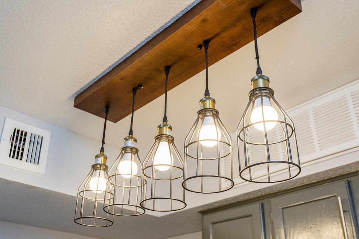A modern ceiling light fixture with five hanging bulbs enclosed in wire cages, mounted on a wooden beam. The fixture adds a stylish touch to the room, with soft light illuminating the space. The background includes a section of a wall with an air vent and hints of cabinetry.