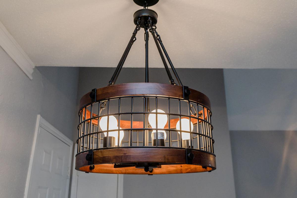 A modern pendant chandelier with a circular wooden frame and metal grid, featuring six exposed light bulbs. The fixture is hung from a ceiling mount, with gray walls in the background, creating a contemporary ambiance.