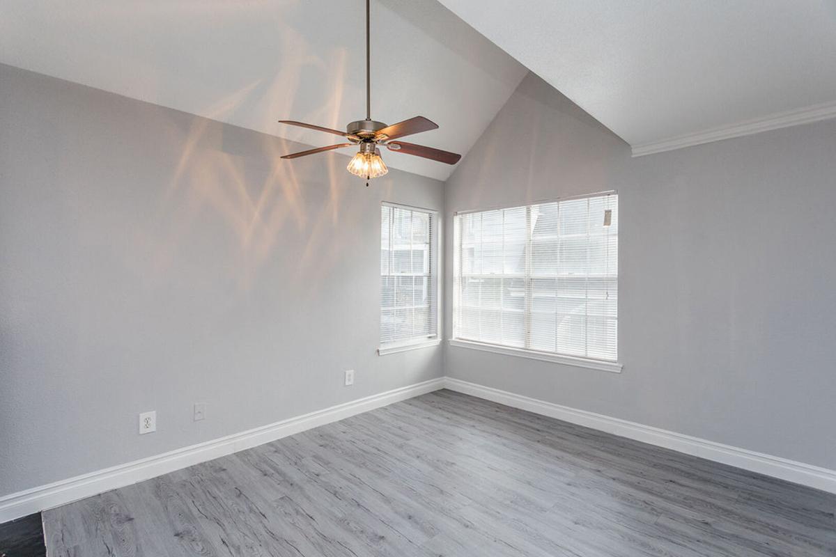 A bright, empty living room featuring a ceiling fan with a light fixture, large windows allowing plenty of natural light, and light gray walls. The floor has a modern wood-like appearance, and the high ceiling adds to the spacious feel of the room.