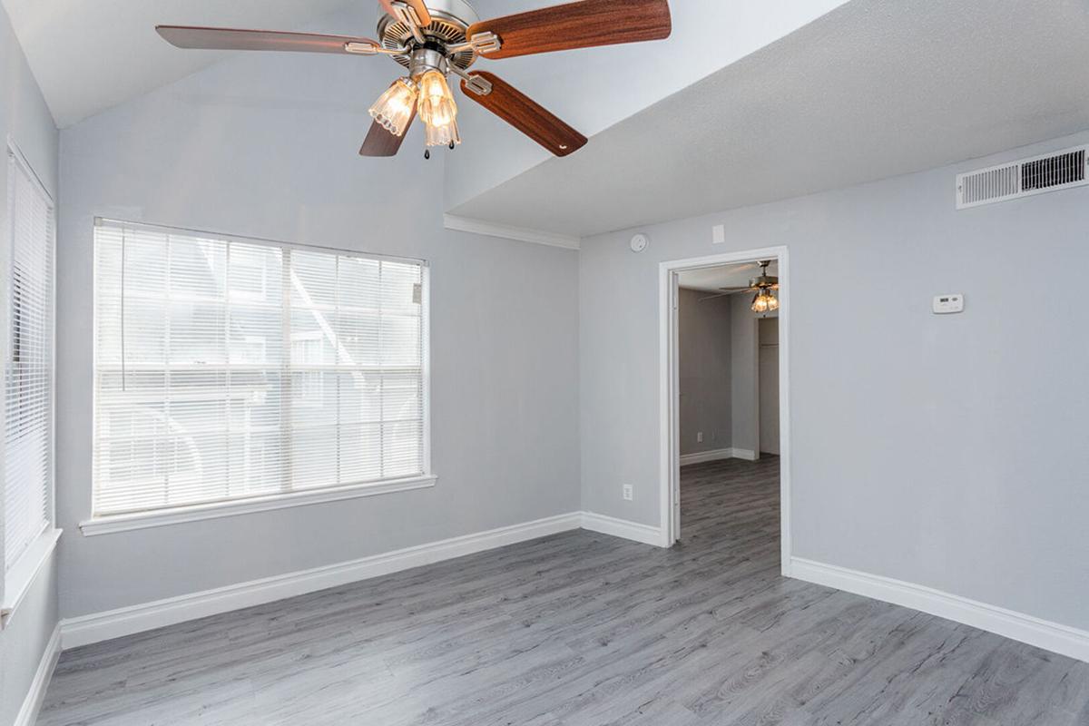 Bright and airy room with light gray walls and wooden flooring. A ceiling fan with light fixtures hangs from a vaulted ceiling. A large window provides natural light, and an open doorway leads to another room. The space is clean and modern, ideal for living or working.