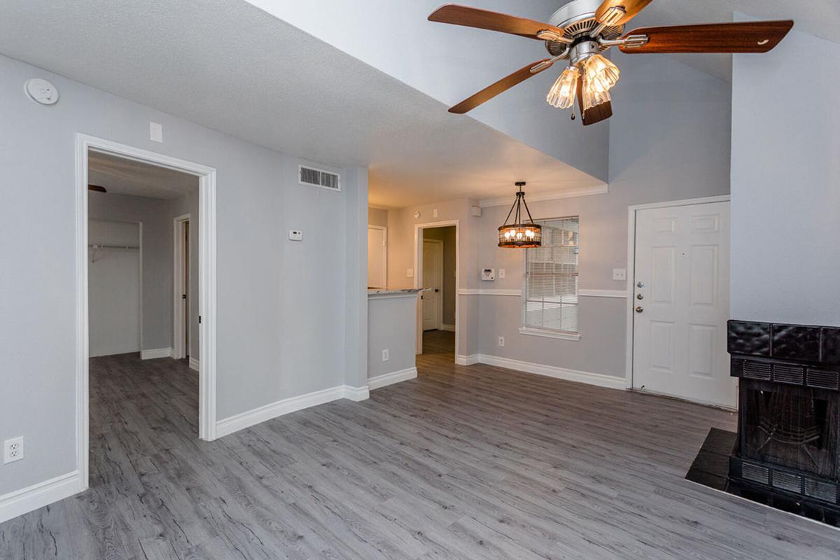 Interior of a modern living room featuring gray walls, wooden flooring, and a ceiling fan with lights. There’s a front door on the right, a fireplace with a black surround, and an open layout leading to another room with a kitchen visible in the background. Ample natural light is coming through the windows.