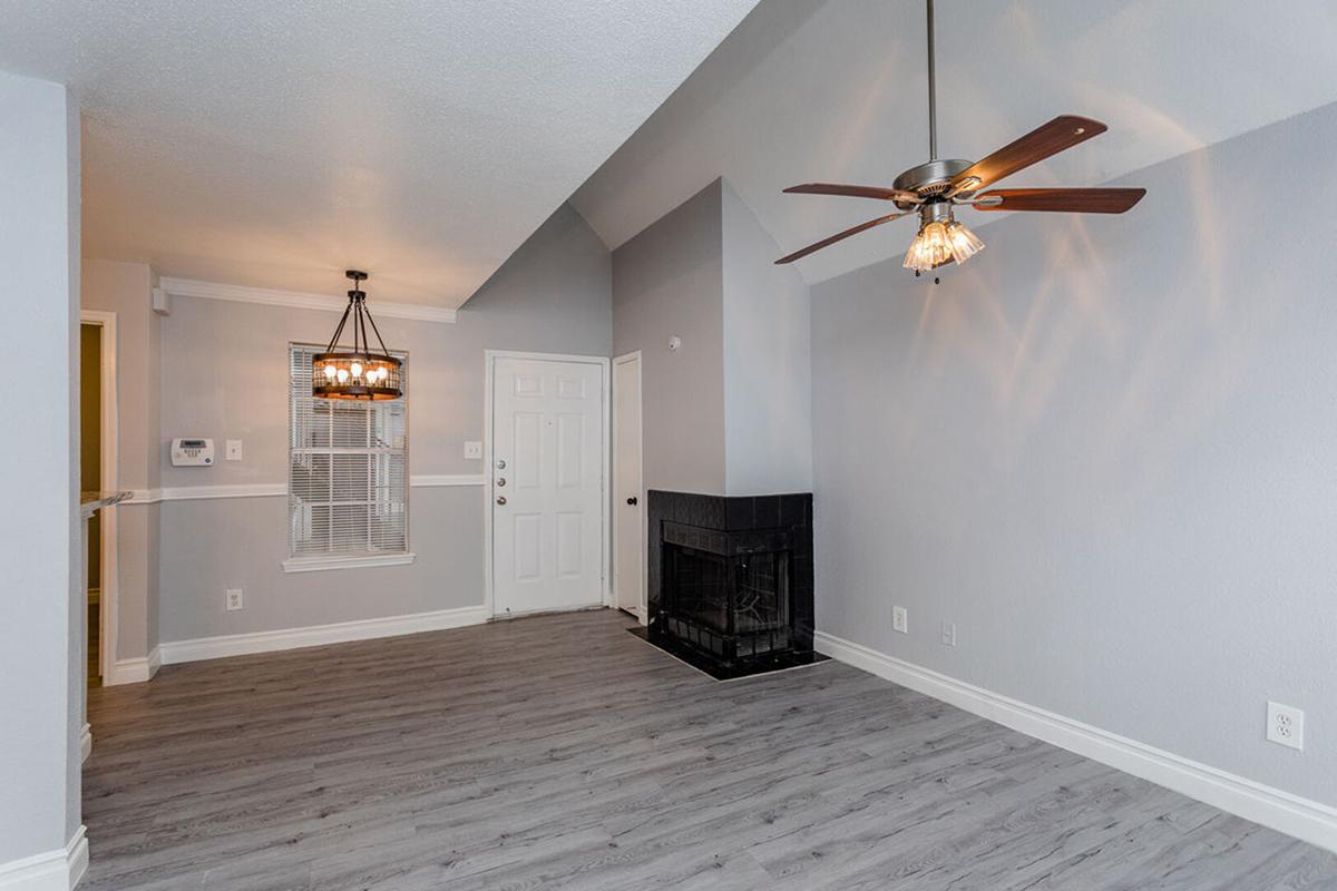 A spacious living room featuring light gray walls, a ceiling fan with a light fixture, and a decorative fireplace. The flooring is a light wood finish, and there is a window allowing natural light. The room has an entrance door and a small chandelier hanging near the ceiling.