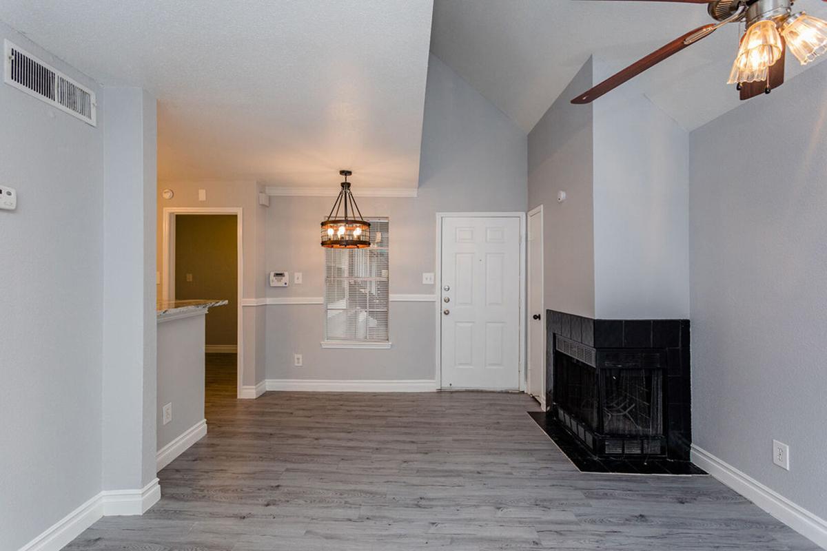 A spacious living room with light gray walls and a vaulted ceiling. It features a ceiling fan, a black tiled fireplace, and a decorative light fixture. A doorway leads to the entrance, while a bar area opens to the kitchen. Natural light enters through the window, enhancing the airy atmosphere.