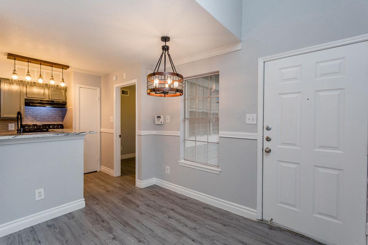 Bright and modern entryway featuring a stylish light fixture, gray walls, and vinyl flooring. On the left, a kitchen area with bar seating is visible. A front door with a vertical window is on the right, and a window with blinds adds natural light to the space.