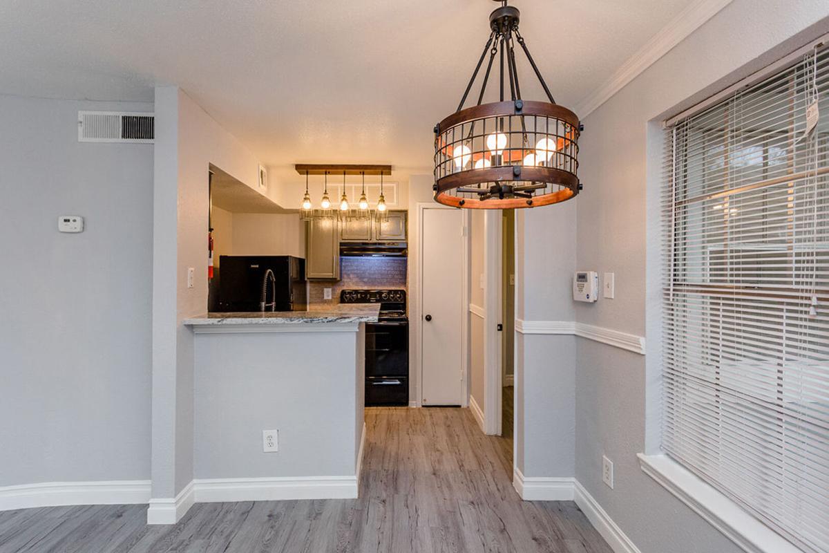 A modern kitchen and dining area with gray walls and wood-like flooring. The kitchen features dark cabinetry and appliances, along with a breakfast bar. A stylish, hanging light fixture adds visual interest, and the space is bright with natural light coming through a window with blinds.