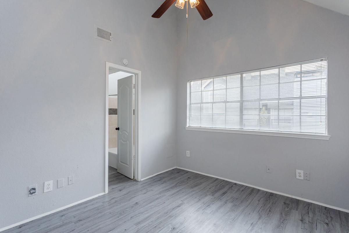 A well-lit room with light gray walls and wood laminate flooring. It features a ceiling fan with wooden blades, a large window with blinds, and a door leading to another space. The overall atmosphere is clean and modern, suitable for various uses such as a bedroom or office.