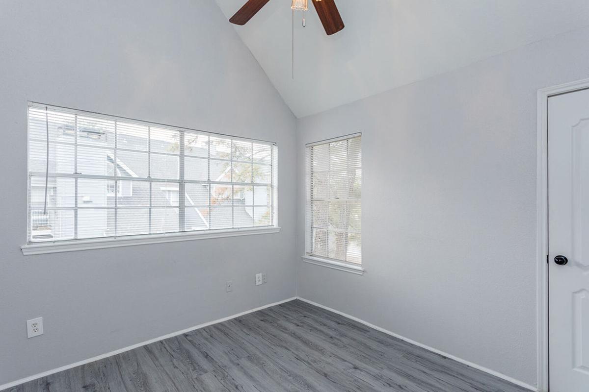 A bright, empty room with light gray walls and wooden flooring. It features a vaulted ceiling with a ceiling fan, large windows with blinds that let in natural light, and a single door on the right side. The space feels clean and open, ideal for various uses such as an office or bedroom.