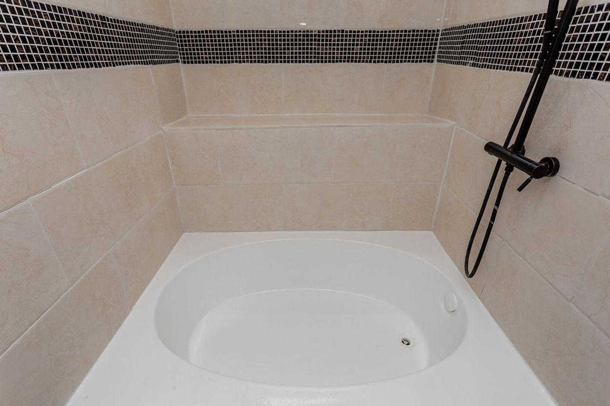 A clean, modern bathroom featuring a white bathtub with a black and white tiled backsplash. The space includes a wall-mounted showerhead and a built-in shelf above the tub, all set against light-colored walls.