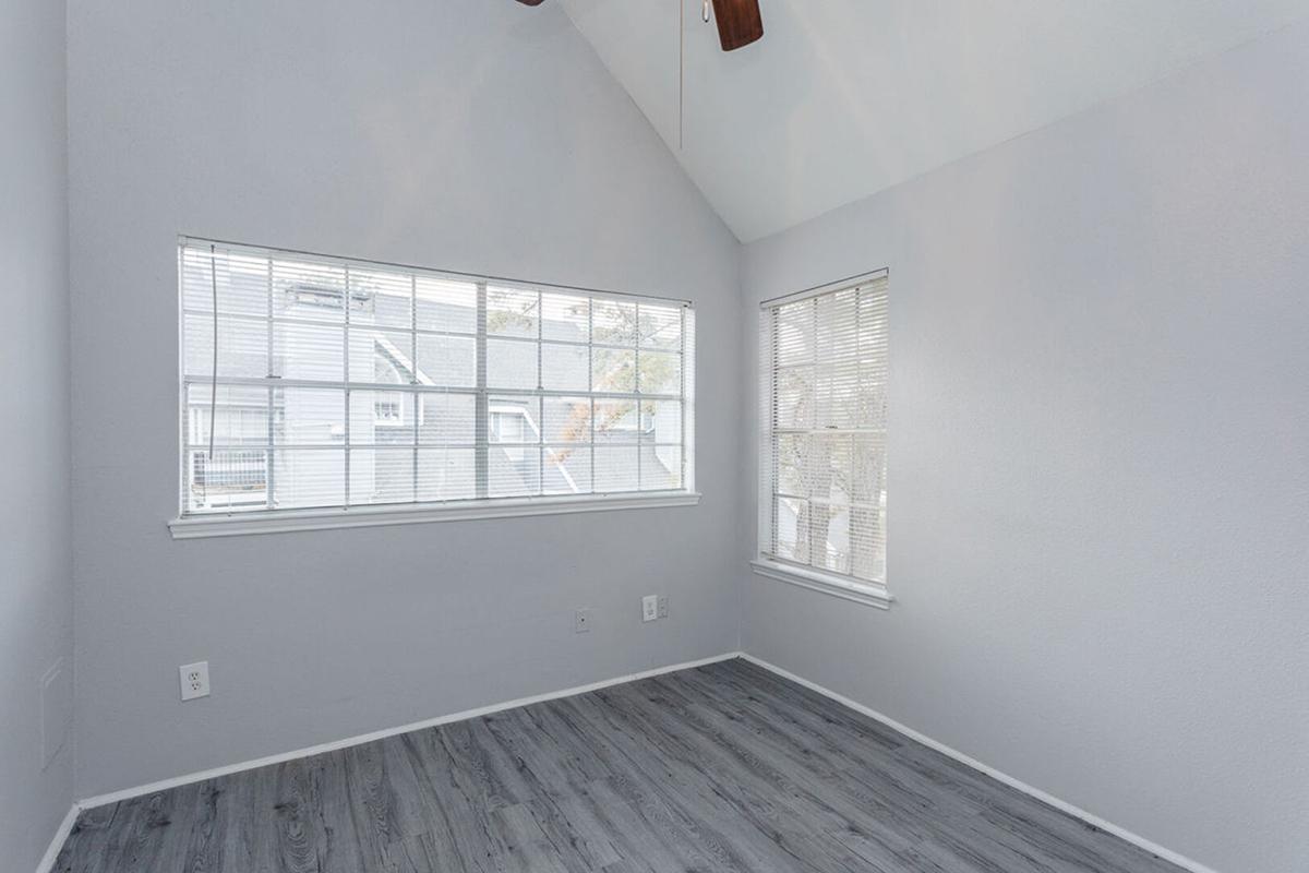 A bright, empty room with light gray walls and a wooden floor. It features a large window with blinds on one wall, allowing natural light to fill the space. The ceiling is high with exposed beams, creating an open and airy feel. The room is uncluttered, suggesting it is ready for new furnishings.