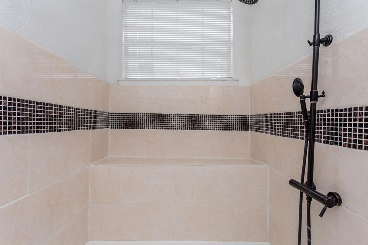 A modern shower space featuring light-colored tile walls with a dark tile accent strip. The shower area includes a showerhead mounted on the wall, a built-in bench, and a window with horizontal blinds providing natural light.