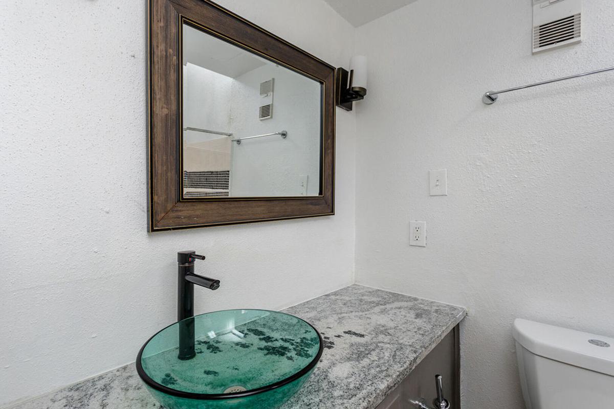 A modern bathroom featuring a glass vessel sink on a granite countertop, a large rectangular mirror with a dark wooden frame, and a wall-mounted light fixture. The walls are painted white, and there is a toilet visible in the background.