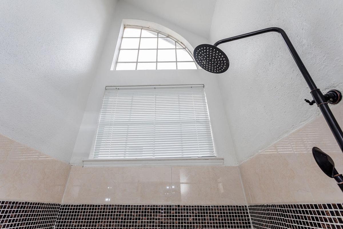 A modern shower with a large, round showerhead. The walls are tiled with a mix of cream and black tiles. Above, there is a large arched window letting in natural light, and a set of white blinds is partially closed. The ceiling is high, featuring a clean white finish.