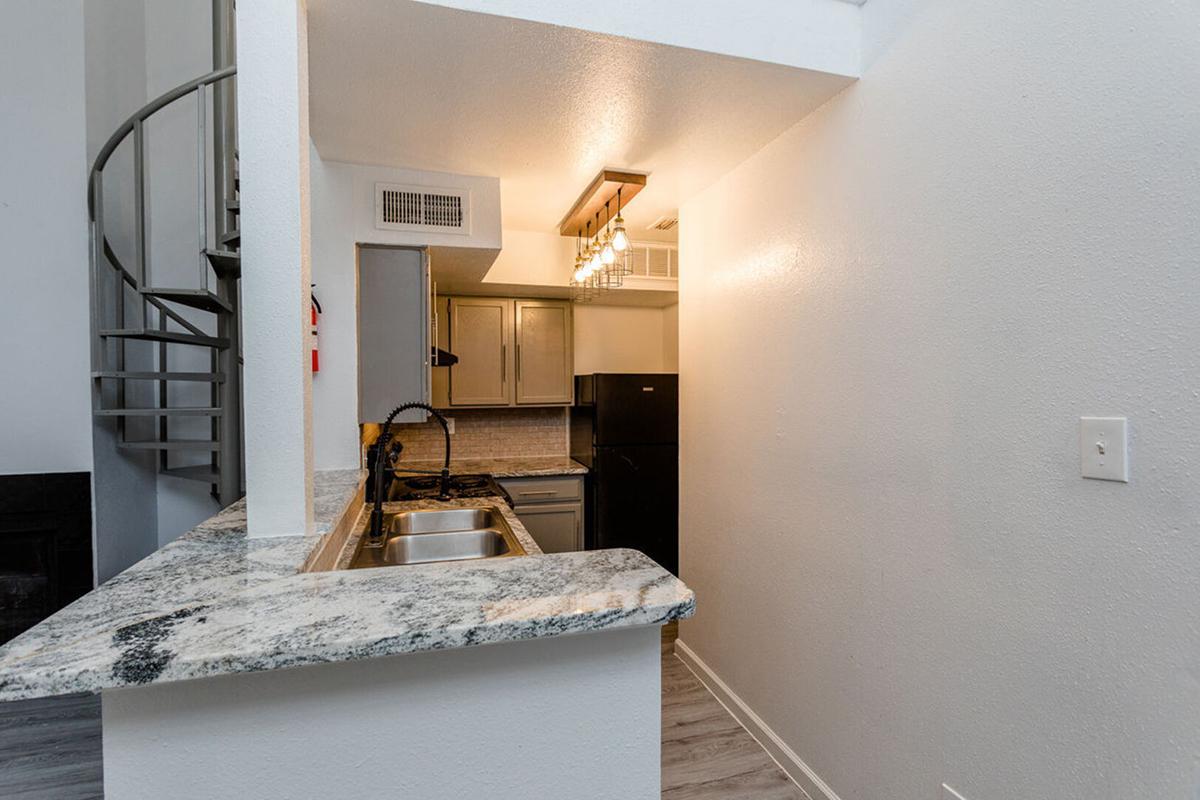 A modern kitchen with a granite countertop, stainless steel sinks, and kitchen appliances including a stove and refrigerator. A spiral staircase is visible in the background, leading to an upper level. The walls are painted white, creating a bright and open atmosphere.