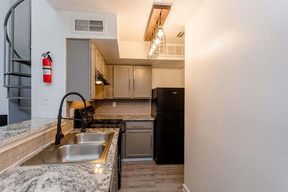 A modern kitchen featuring gray cabinetry, stainless steel appliances, and a double sink with a modern faucet. The space includes a black refrigerator, an oven, and a range above the stove. Natural light comes in through a small window, and a spiral staircase is visible in the background. There is a fire extinguisher mounted on the wall.