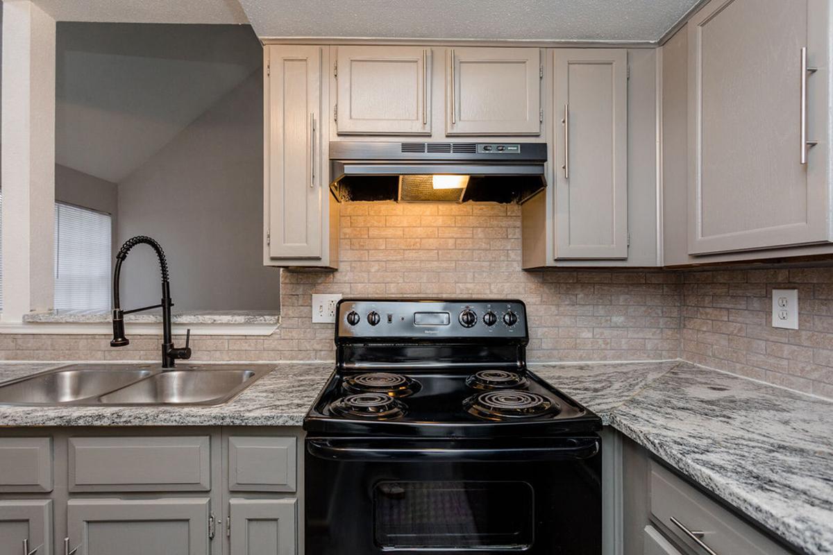 A modern kitchen featuring a black electric stove, light gray cabinets, and a dual-basin sink. The countertop is made of a marble-like material, and there's a vent hood above the stove. The backsplash consists of light-colored bricks, creating a contemporary aesthetic.
