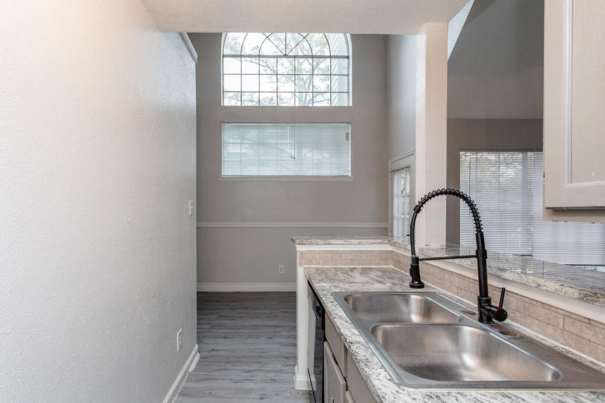 A modern kitchen featuring a double sink with a black faucet, light-colored cabinets, and granite countertops. The space is illuminated by natural light coming through a large window above the sink. The flooring is a light gray laminate, and there are additional windows allowing for ample brightness in the area.