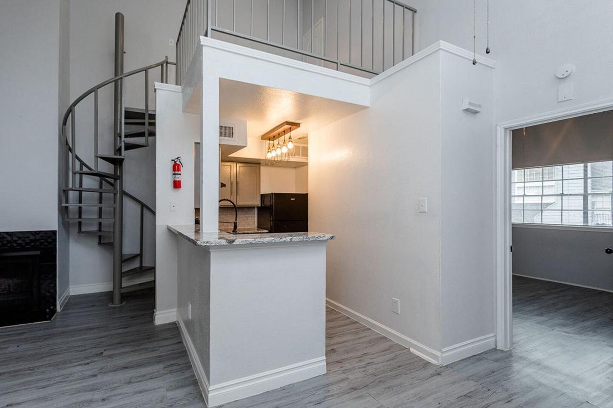 Interior view of a modern apartment featuring a spiral staircase. The kitchen area has a granite counter with pendant lighting and dark cabinetry. Light-colored walls and flooring create a spacious feel, with large windows allowing natural light to fill the space. A fire extinguisher is mounted on the wall.