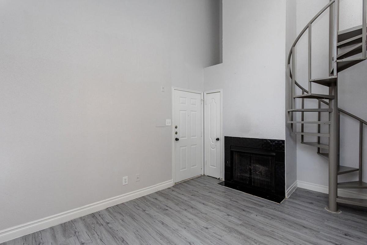 A spacious room with light gray walls and wood-style flooring. The interior features a sleek black fireplace, a white front door, and a modern spiral staircase leading to an upper level. The ceiling is high, creating an open and airy feel in the space.