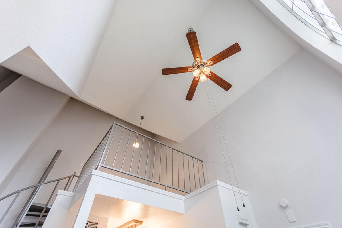 Interior view of a multi-level space featuring a modern ceiling fan with wooden blades, high white walls, and a spiral staircase. Natural light filters in through a large window, enhancing the open and airy atmosphere. The design combines contemporary elements with a spacious feel.