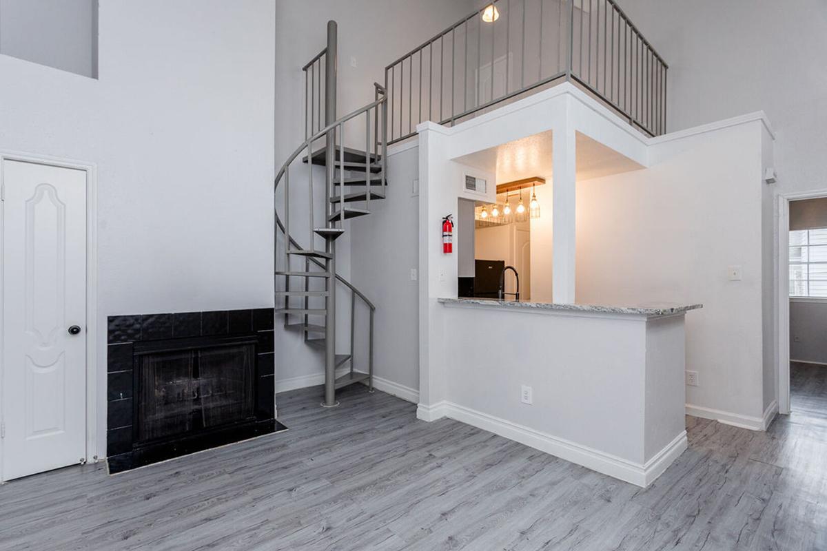 Modern interior of a loft-style living space featuring a spiral staircase, a fireplace, and an open kitchen area with a breakfast bar. The walls are painted light colors, and the flooring is a light gray wood finish. Large windows provide natural light to the room.