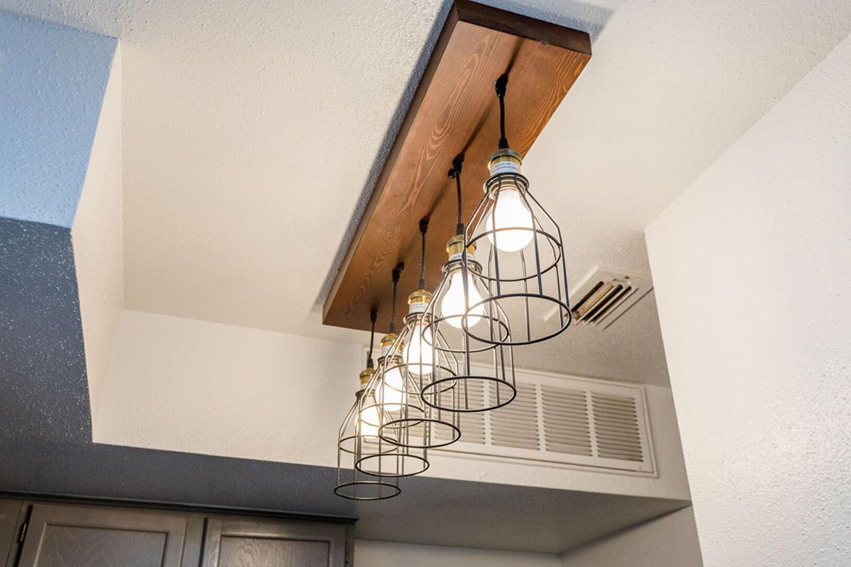 Five pendant lights with clear bulbs are suspended from a wooden beam attached to the ceiling. The lights are enclosed in black wire cages, creating a modern industrial look. The ceiling has a textured finish, and there is a vent in the background. The scene is bright and stylish, ideal for a kitchen or dining area.