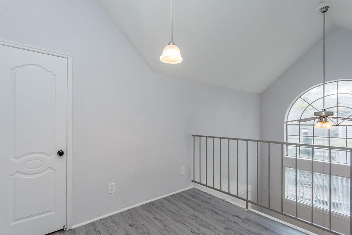 A bright, airy loft space with high ceilings and pale gray walls. A light fixture hangs from the ceiling, and a railing separates the loft area from a lower level. A large arched window allows natural light to flood in, while a closed door is visible on the left side of the image.
