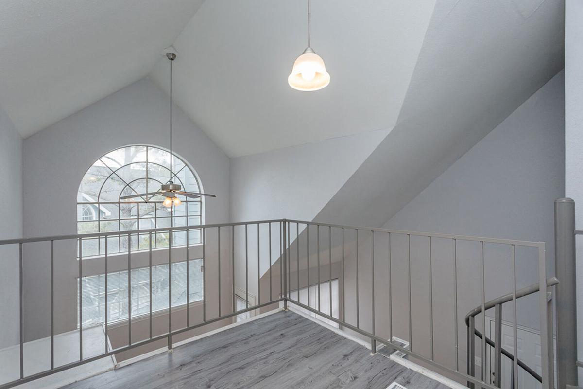 Interior view of a light-filled loft area with a high ceiling and large arched window. A ceiling fan hangs from the ceiling, and a metal railing surrounds the loft space. A staircase curves downwards in the background, and the walls are painted in soft gray tones.