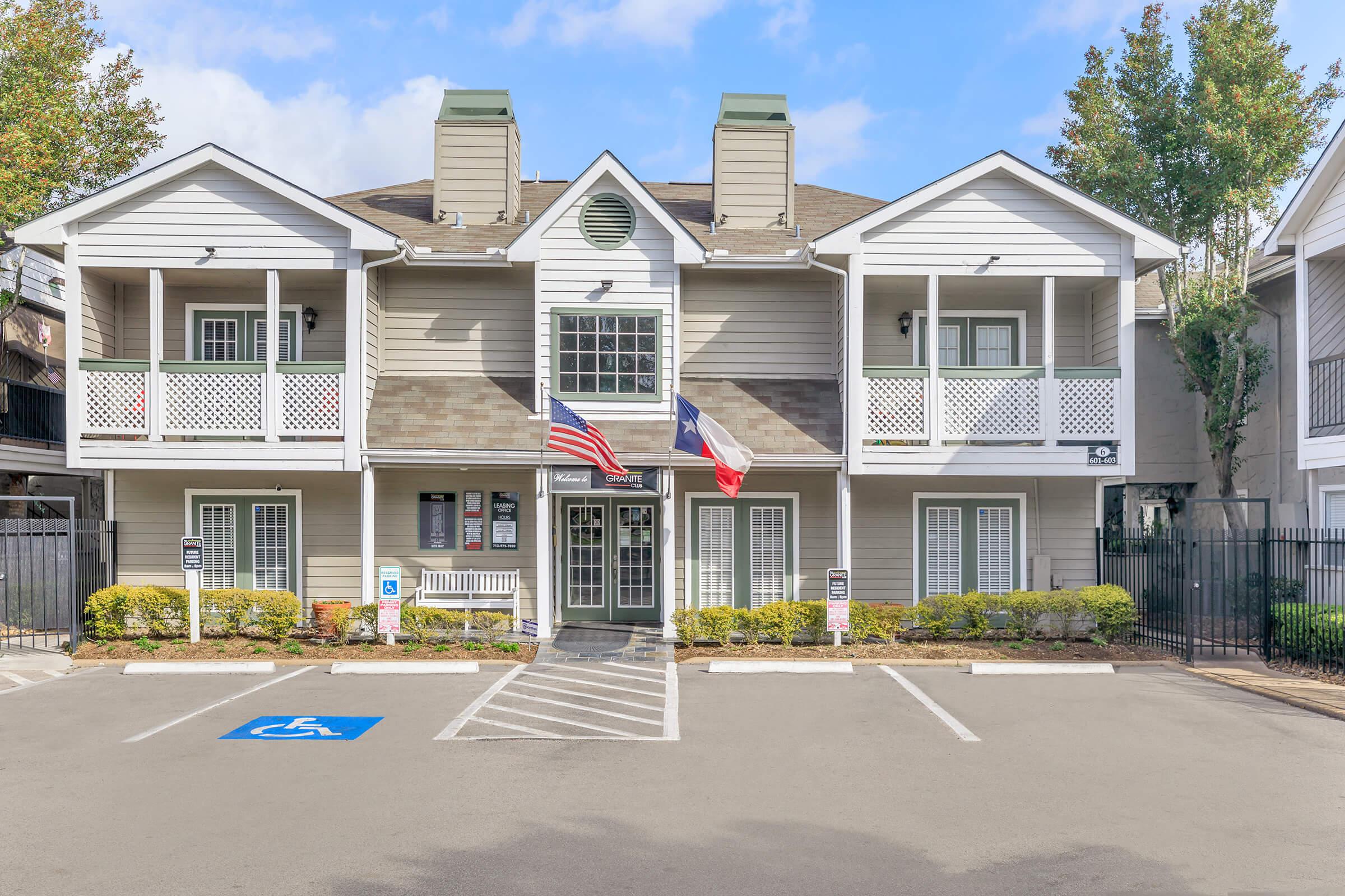 A two-story building with a light gray exterior, featuring balconies with decorative railings. The main entrance has American and Texas flags displayed. The surrounding area includes well-maintained landscaping and a designated handicapped parking space in front. Bright, clear skies are visible above.