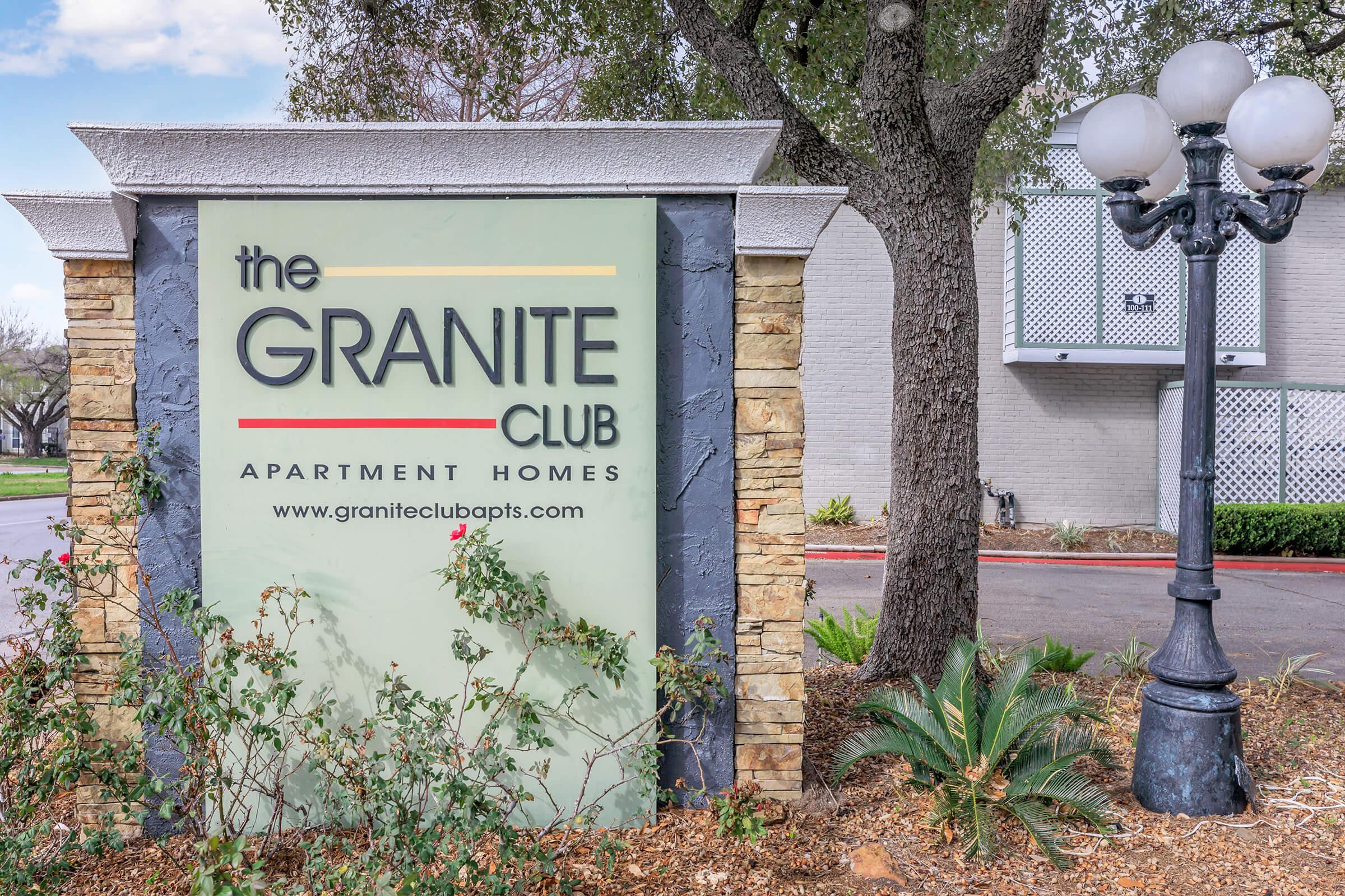 Sign for "the GRANITE CLUB" apartment homes, featuring a stone and modern design with green foliage surrounding it. A lamp post is visible nearby, suggesting a welcoming entrance to the apartment community. The setting appears well-maintained and inviting.
