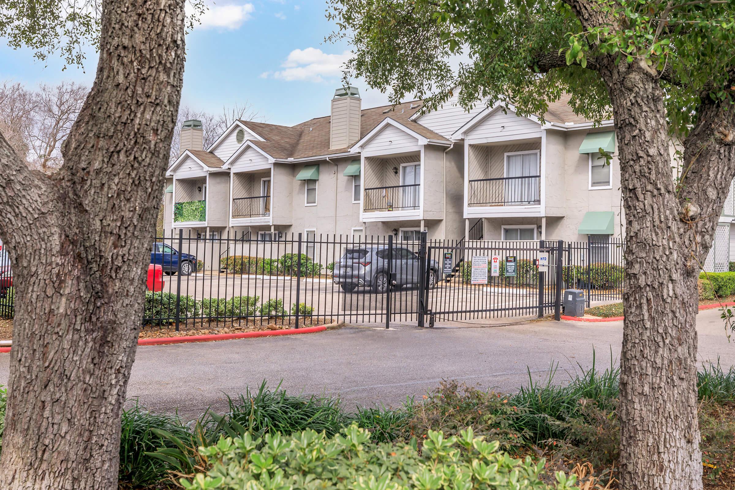 A view of a gated apartment complex with two-story buildings, featuring balconies and green accents. The area is surrounded by trees and shrubs, with a parking lot visible in front. Clear blue sky and a few clouds in the background enhance the scene.
