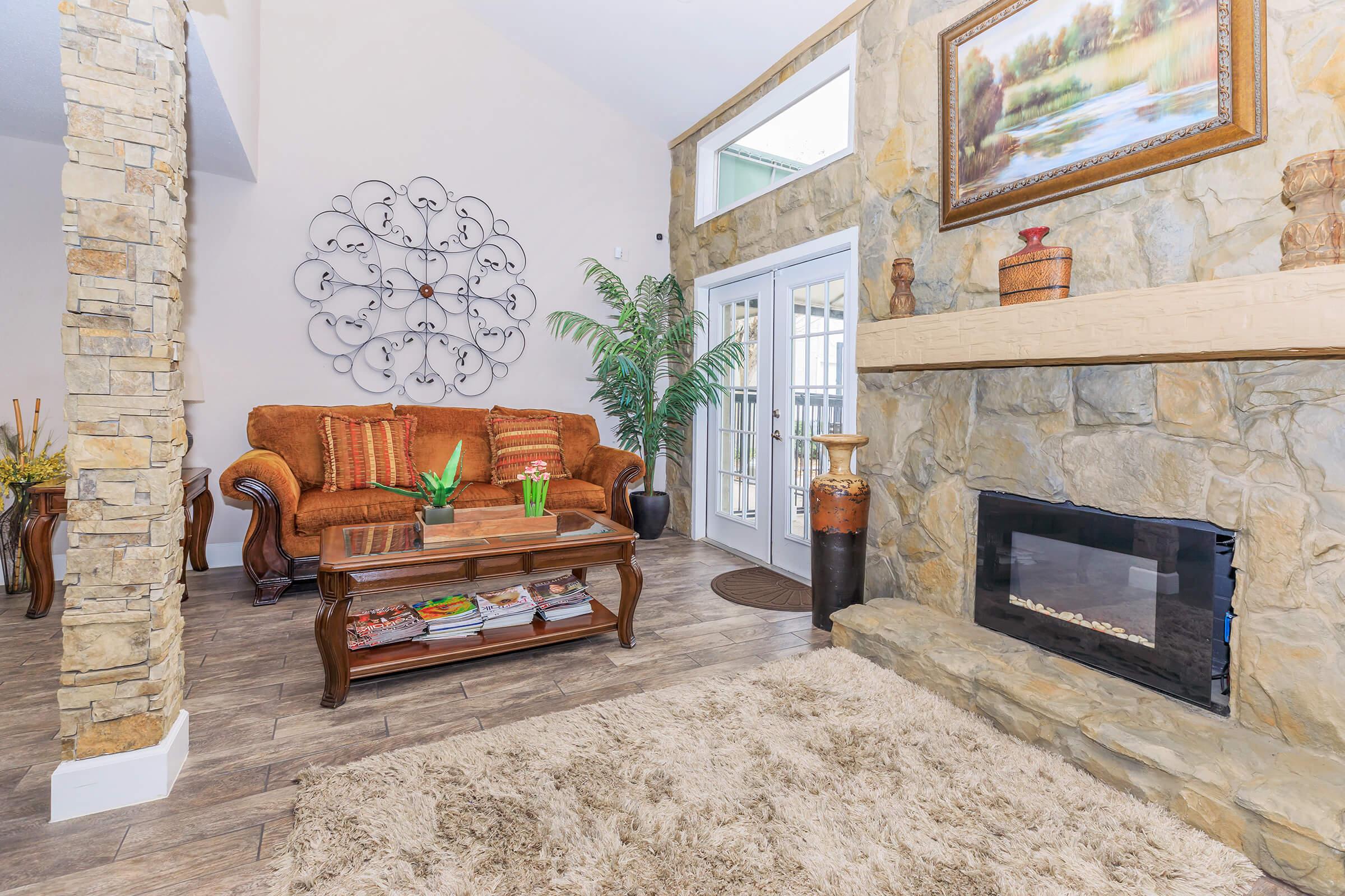 Cozy living room featuring a brown sofa with decorative cushions, a stone fireplace, and a stylish coffee table with magazines. A large wall décor adorns one side, while a green plant adds a touch of nature. Sunlight streams in through glass doors leading to an outdoor area, with a soft area rug on the floor.